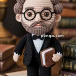 Crocheted Machado de Assis doll wearing glasses and a suit holding a book, placed on an open book with a pen, surrounded by more books and a lamp in the background.