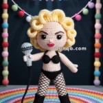 A crocheted doll with curly yellow hair, wearing black lingerie and fishnet stockings, holds a microphone. It stands on a colorful crocheted rug with a backdrop that reads "LIKE A PRAYER" in multicolored letters.