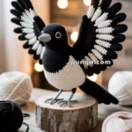 A crocheted magpie with black and white wings is perched on a wooden stand, surrounded by yarn balls and crochet hooks, with soft fairy lights in the background.