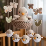 A handmade crocheted mobile featuring a large bird in a nest at the center, surrounded by smaller birds hatching from eggs, butterflies, and leaves, hanging over a crib.