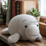 A knitted manatee plush toy on a wooden shelf surrounded by books and a few potted succulents.
