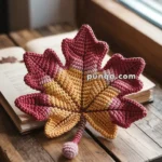 A crocheted maple leaf with red, yellow, and orange hues lies on a wooden table next to an open book by a window.