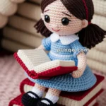 A crocheted doll with brown hair and pigtails, wearing a blue dress and black shoes, is sitting on two crocheted books while holding an open crocheted book.