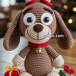 A crocheted dog with reindeer antlers, sitting on a white knitted mat with small gift boxes beside it, adorned with a red collar featuring a bell, is placed against a blurred cozy indoor background with lights.
