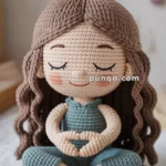 A crochet doll with brown hair and a bun, seated cross-legged on a round mat, wearing a blue outfit and making a heart shape with its hands.