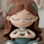 A crochet doll with brown hair and blue clothing sits cross-legged on a braided mat, forming a heart shape with its hands and smiling with closed eyes.