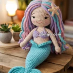 A crocheted mermaid doll with pastel purple and blue hair, wearing a purple top and turquoise tail with a starfish attached to the hair, sitting on a wooden table near small potted plants and a lit lamp.
