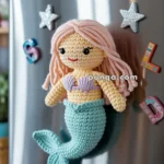 Crocheted mermaid magnet with pink hair and a lavender top attached to a silver refrigerator door, surrounded by star-shaped and letter magnets.