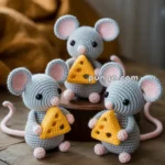 Three crocheted gray mice with pink ears and feet, each holding a small triangular piece of crocheted cheese, are placed on a wooden surface with a brown fabric in the background.