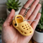 A hand holding a keychain that features a small, yellow, crochet sandal resembling a clog with a strap and decorative holes.