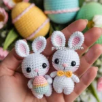 Two crocheted white bunny dolls with pink ears and black eyes, one holding a colorful Easter egg and the other wearing a yellow bow tie, held in a person's hand with pastel-colored Easter eggs and flowers in the background.