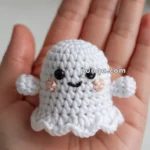A small crocheted ghost with a cute smiley face and pink cheeks, held in the palm of a hand.