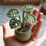 Crocheted Monstera plant placed in a small crocheted pot, held in a person's hand, with a blurred background showing a wooden box labeled "PLANT LOVE" and other succulents.