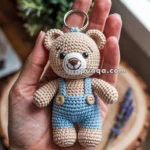 A hand is holding a crocheted teddy bear keychain wearing blue overalls, with a plant and window in the blurred background.
