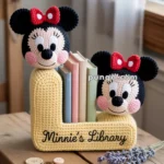 Crocheted bookends featuring two Mickey Mouse-inspired faces with red bows, labeled "Minnie's Library," holding books on a wooden table with buttons and a sprig of lavender nearby.