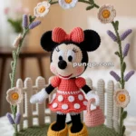 Crocheted doll resembling a cartoon character with round ears, wearing a red polka-dot dress and red bow, standing on a green base with crocheted flowers and a white picket fence.