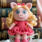 A crocheted doll resembling a pig with blonde hair and a pink dress, sitting in a floral teacup labeled "Miss Piggy" in front of a stack of books, with a red rose in the background.