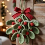 A crocheted mistletoe with green leaves and red berries, adorned with a red bow, hanging on a wooden door handle in a festive setting with blurred lights in the background.