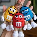 Crocheted keychains of M&M's characters in yellow, red, and blue, each featuring cartoon faces, held in a person's hand with a background of a blurred room.