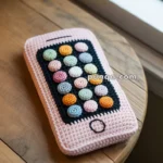 A crochet phone case designed to look like a smartphone, featuring colorful crochet app icons on a black screen background, set on a wooden table by a window.