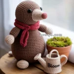 A crocheted mole toy wearing a red scarf is sitting on a wooden stool beside a potted plant and a small crocheted watering can labeled "Bloom."