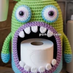 A crochet monster design with large eyes and a wide open mouth that holds a roll of toilet paper, placed on a bathroom countertop.