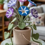 A crocheted potted plant featuring colorful flowers with white star patterns, green stems, and heart-shaped leaves, placed on a wooden table with a tag reading 'Handmade with love.'