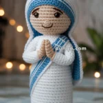 A crocheted doll with a smiling face and hands together in a praying position, dressed in a white robe with blue trim and a matching headscarf, standing on a wooden surface with blurred lights in the background.