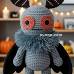 A crocheted plush toy resembling a moth with large orange eyes, black wings, and segmented ears, standing on a wooden surface with Halloween decorations in the background.