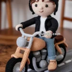 A crocheted doll wearing a black jacket and goggles sits on a crocheted motorcycle, placed on a crocheted road surface, with a cozy indoor setting.