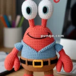 A crocheted toy crab with large eyes, wearing a blue shirt and brown shorts with a belt, placed on a wooden desk.