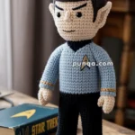 A crocheted doll resembling a well-known science fiction character, dressed in a blue uniform with a gold insignia, stands on a wooden table next to a book titled "Star Trek."