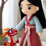 A crocheted doll of a young woman with long black hair in traditional attire, standing on a green mat and holding hands with a small crocheted dragon. Cherry blossoms are visible in the background.