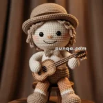 A crocheted musician doll sits on a wooden stool, holding a small guitar. The doll is dressed in a beige top hat, bow tie, and shorts, with a cheerful smile and rosy cheeks. It has curly yarn hair and wears matching shoes and socks. The background is softly lit with brown drapes.