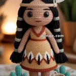 A handmade crochet doll with braided black hair, wearing a tan dress decorated with black and red patterns and two white feathers in her headband. The doll stands on a crocheted orange mat, accompanied by small crocheted cactus plants and a basket.