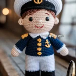 A crocheted doll dressed in a navy uniform with gold anchor and buttons, complete with a sailor hat, standing next to an antique pocket watch on a wooden surface.
