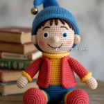 A crocheted doll with a blue hat adorned with a bell, featuring brown hair, blue eyes, and a cheerful expression. The doll wears a red jacket, yellow shirt, and blue pants with yellow cuffs, seated on a wooden table next to a stack of books.