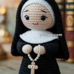 A crocheted doll of a nun in a black habit, holding a cross with a beaded chain, placed on a wooden surface with books in the background.
