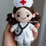 A hand holding a crocheted doll dressed as a nurse, wearing a white outfit with a red cross on the hat and a stethoscope around the neck.