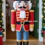 A crocheted nutcracker soldier with a red jacket, blue pants, and a tall black hat stands on a circular base, surrounded by Christmas decorations.