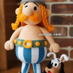 A crocheted doll of a character with a horned helmet, orange mustache, and striped pants, standing next to a small crocheted dog with floppy ears, set against a brick wall background.