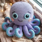 A cute purple crocheted octopus with blue and turquoise-tipped tentacles, large eyes, and a smiling face sits on a knitted surface beside a wooden plaque that reads "Dream Big."