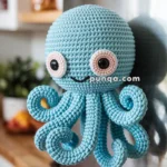 A crocheted blue octopus with cute large eyes and a smile is attached to a stainless steel refrigerator door, with a kitchen setting in the background.