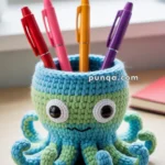 A crocheted octopus-shaped pen holder with large eyes, holding colorful pens on a desk near a window.