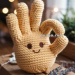 A crocheted plush toy shaped like a hand making an "OK" gesture, with a smiling face embroidered on it, sitting on a wooden surface with blurred lights in the background.