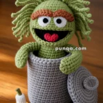 A crocheted green puppet with bushy hair and eyebrows is popping out of a gray crocheted trash can, with a crocheted banana peel and apple core beside it on a wooden surface.