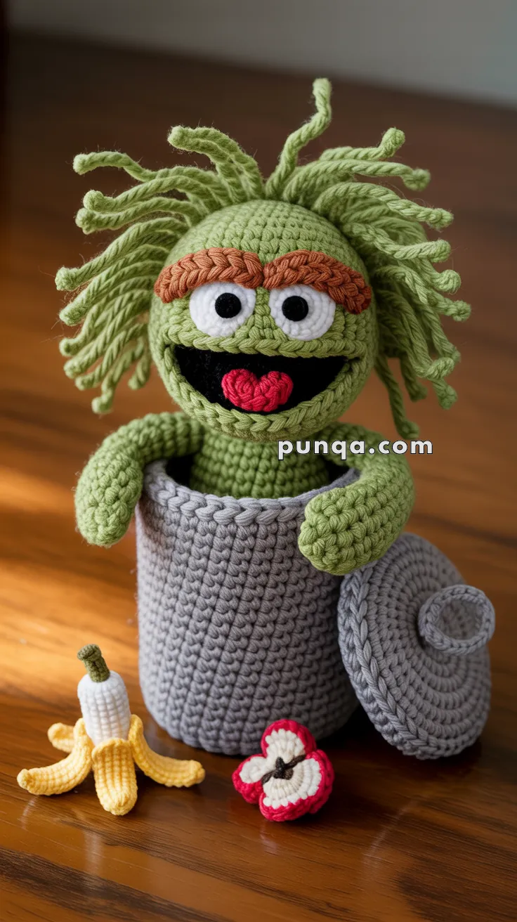 A crocheted green puppet with bushy hair and eyebrows is popping out of a gray crocheted trash can, with a crocheted banana peel and apple core beside it on a wooden surface.