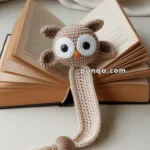 An open book with a crocheted owl bookmark featuring large eyes, an orange beak, and a tassel at the end.