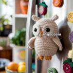 A crocheted owl magnet attached to a refrigerator surrounded by other colorful crocheted fruit magnets, with a bowl of oranges and lemons in the background.