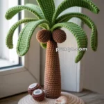 A crocheted palm tree with brown coconuts and green leaves on a circular beige base, placed on a wooden windowsill.